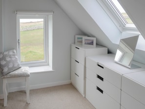 Une pièce avec un plafond en pente, une fenêtre donnant sur l’extérieur, un bureau blanc avec des tiroirs et une chaise avec un coussin.