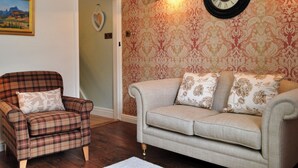 Ein Wohnzimmer mit einem Karoarmlehnstuhl, einem beigen Sofa mit floralen Kissen, einer Wanduhr und einem gerahmten Landschaftsbild.