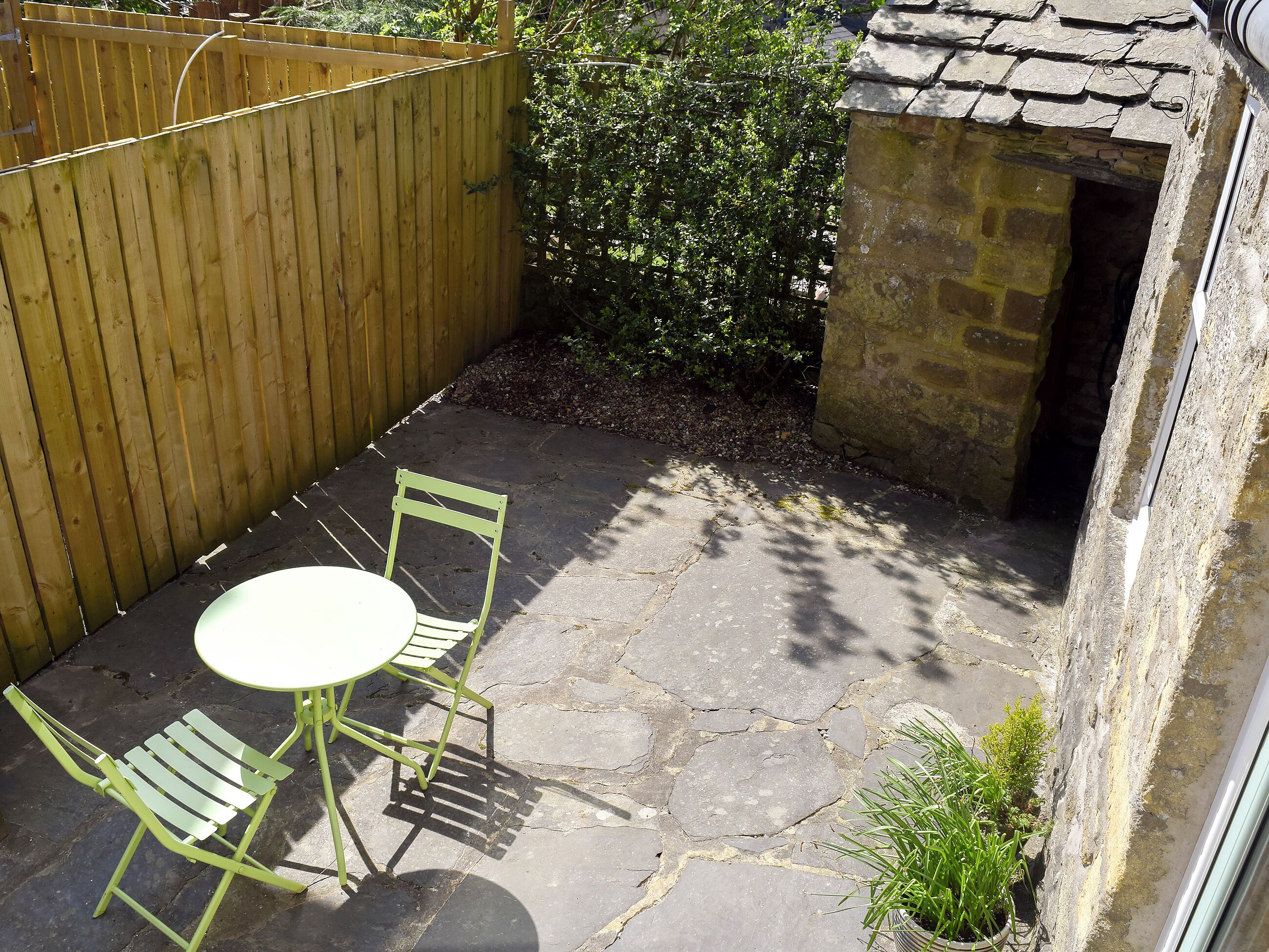 A small patio area with a stone floor, a wooden fence, a green metal chair, a white table, and a small potted plant.