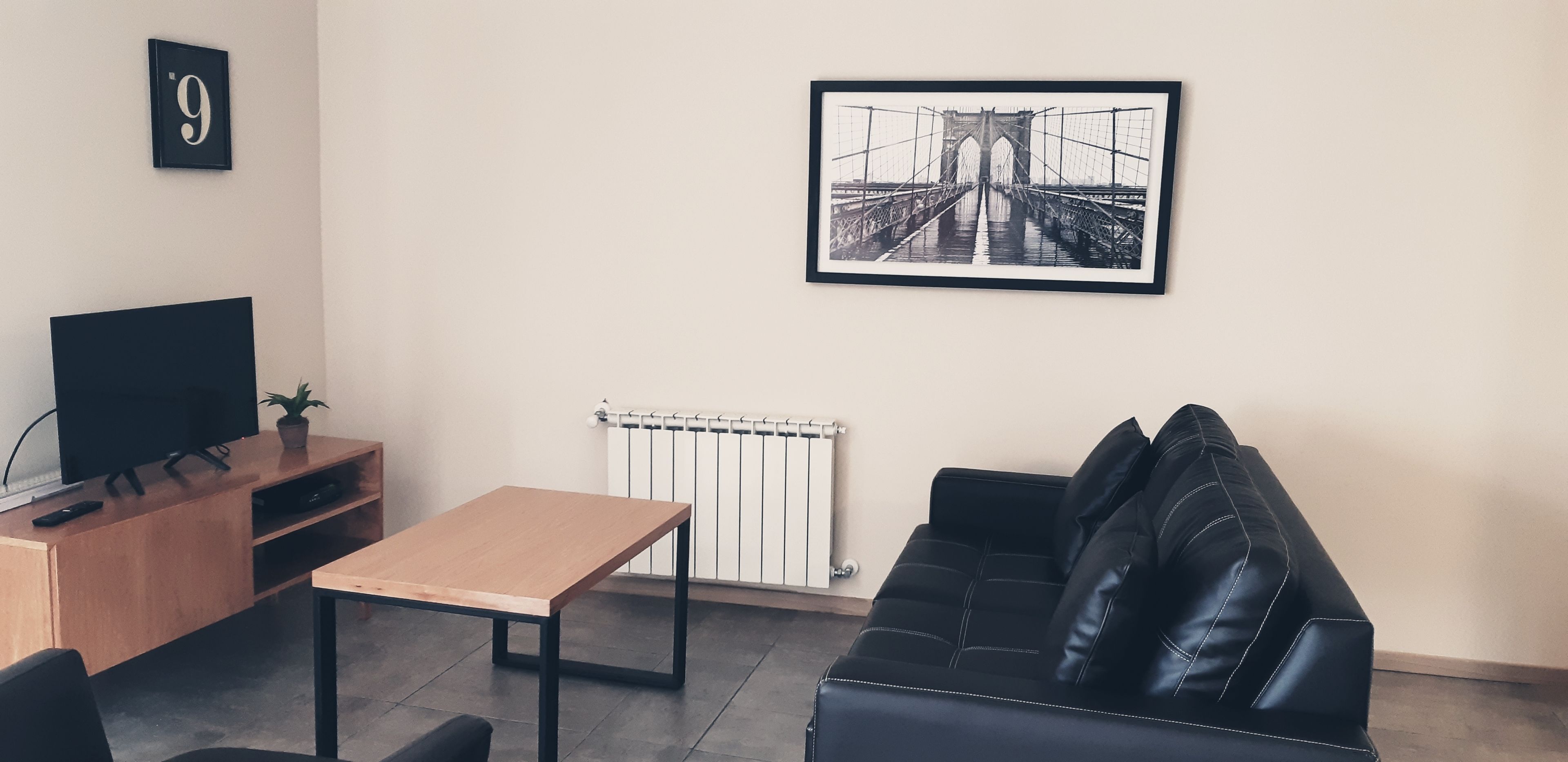 A modern living room with a black leather sofa, a wooden coffee table, a TV on a wooden stand, and a framed picture of a bridge on the wall.