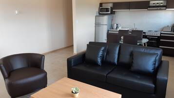 A modern living room with a black leather sofa, a wooden coffee table with a plant, and a kitchen in the background featuring a microwave, refrigerator, and stove.