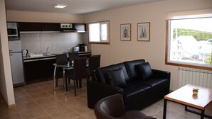 A compact living space with a kitchenette, a black leather sofa, a dining table, and a window offering a view of buildings. - Complejo los Guindos - Ushuaia Flat (Ushuaia)