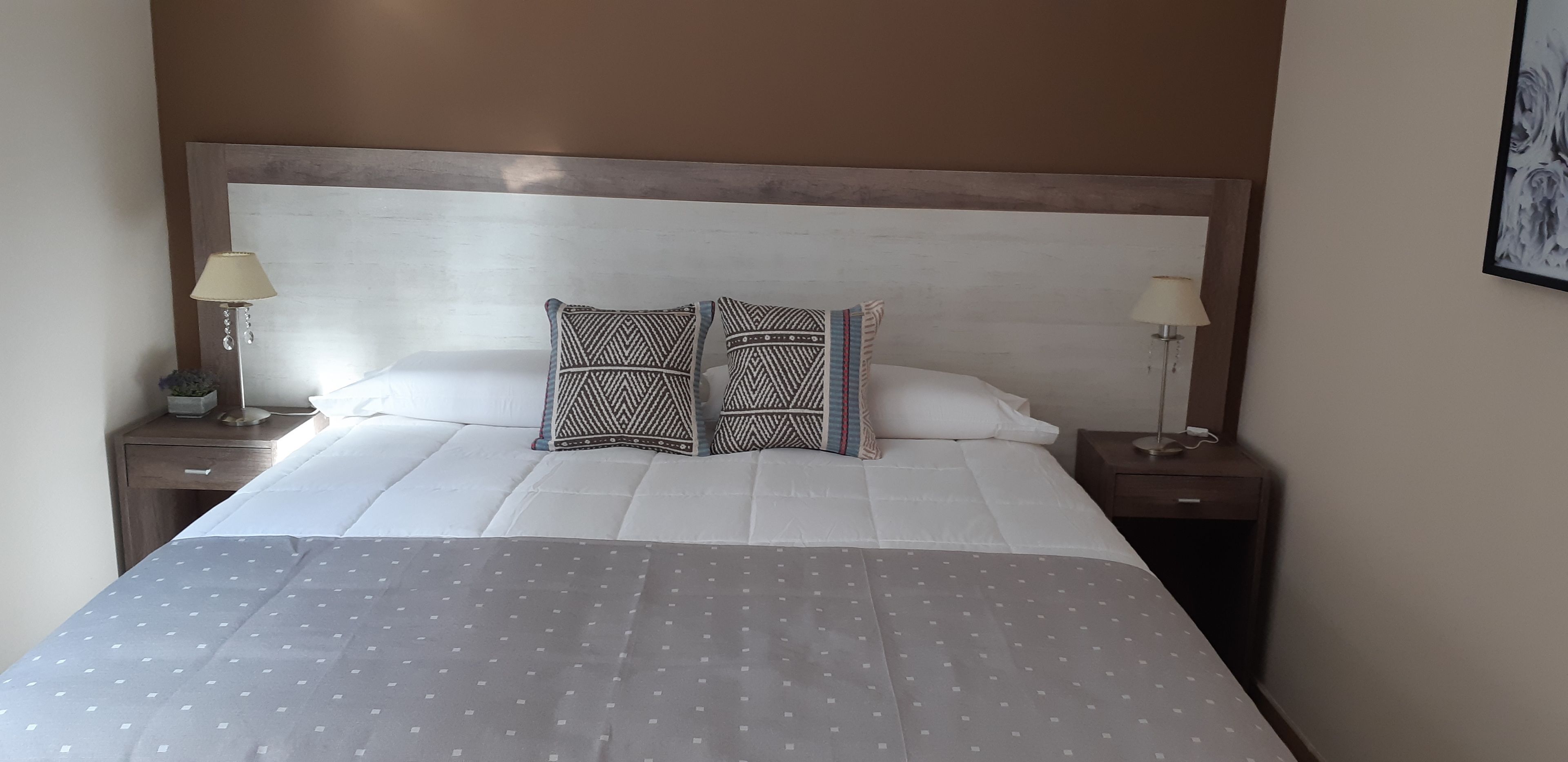 Una cama bien hecha con almohadones estampados, una colcha gris con lunares y dos lámparas de noche sobre mesitas de madera.