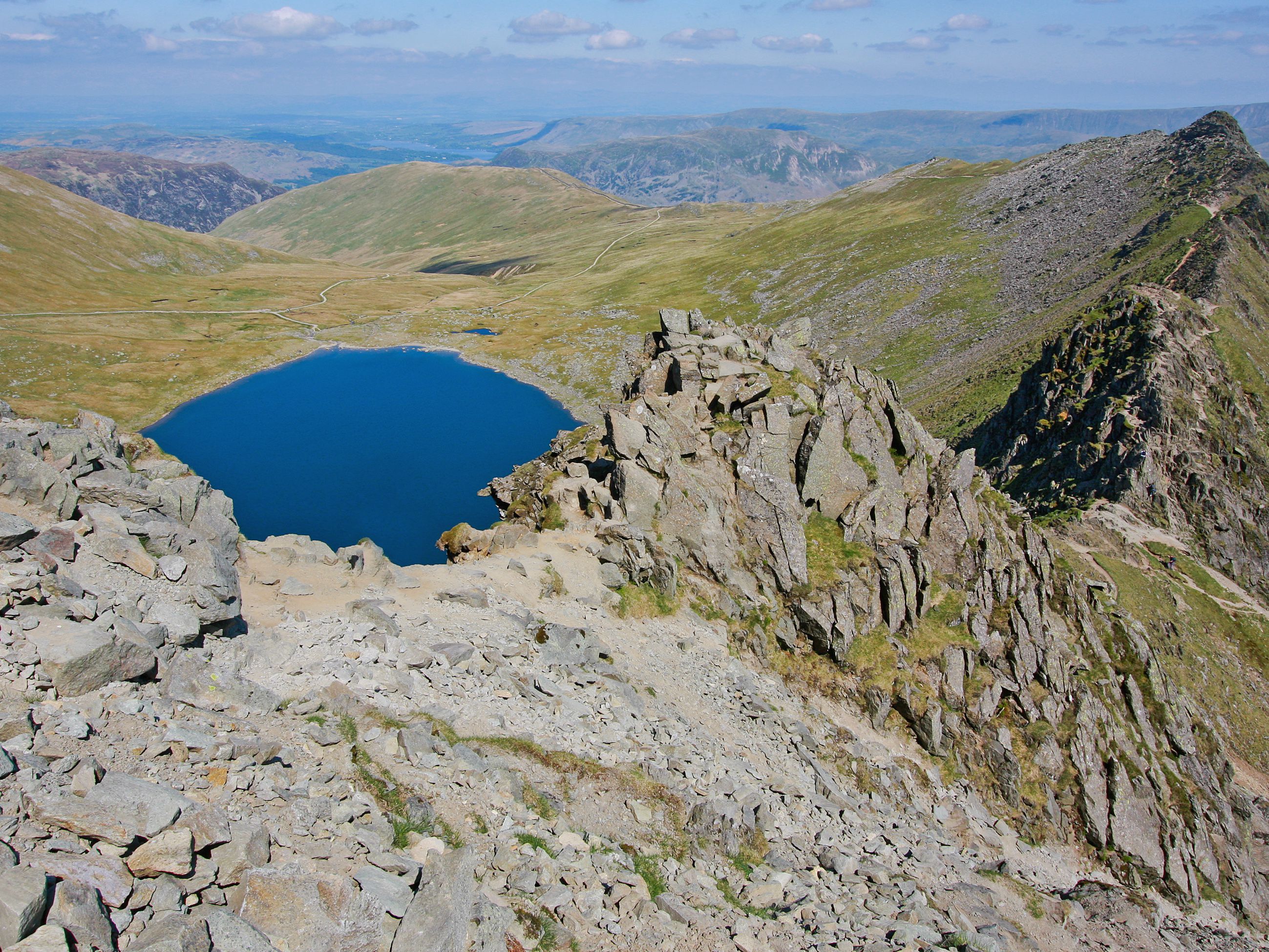 A rugged mountain landscape with a clear blue lake surrounded by rocky terrain and distant hills.