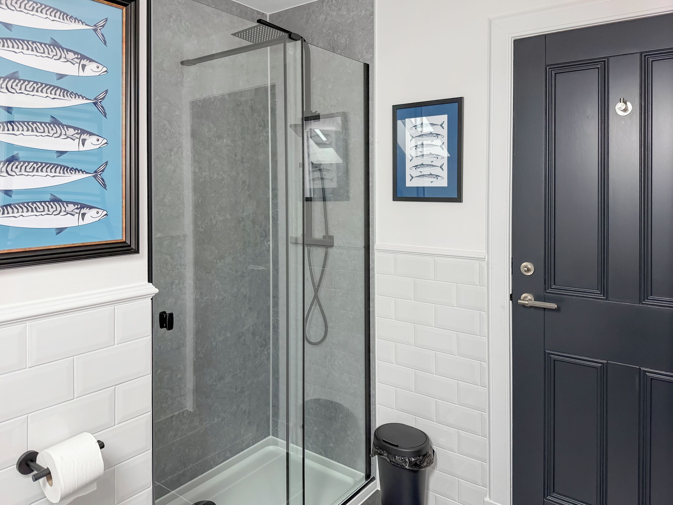 A bathroom with a glass-enclosed shower, a toilet paper holder, a framed fish illustration, and a dark door.