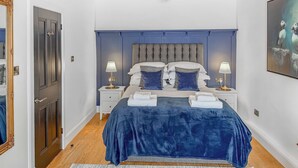 Un dormitorio con una cama grande, dos mesitas de noche con lámparas, un cabecero azul oscuro y un cuadro en la pared.