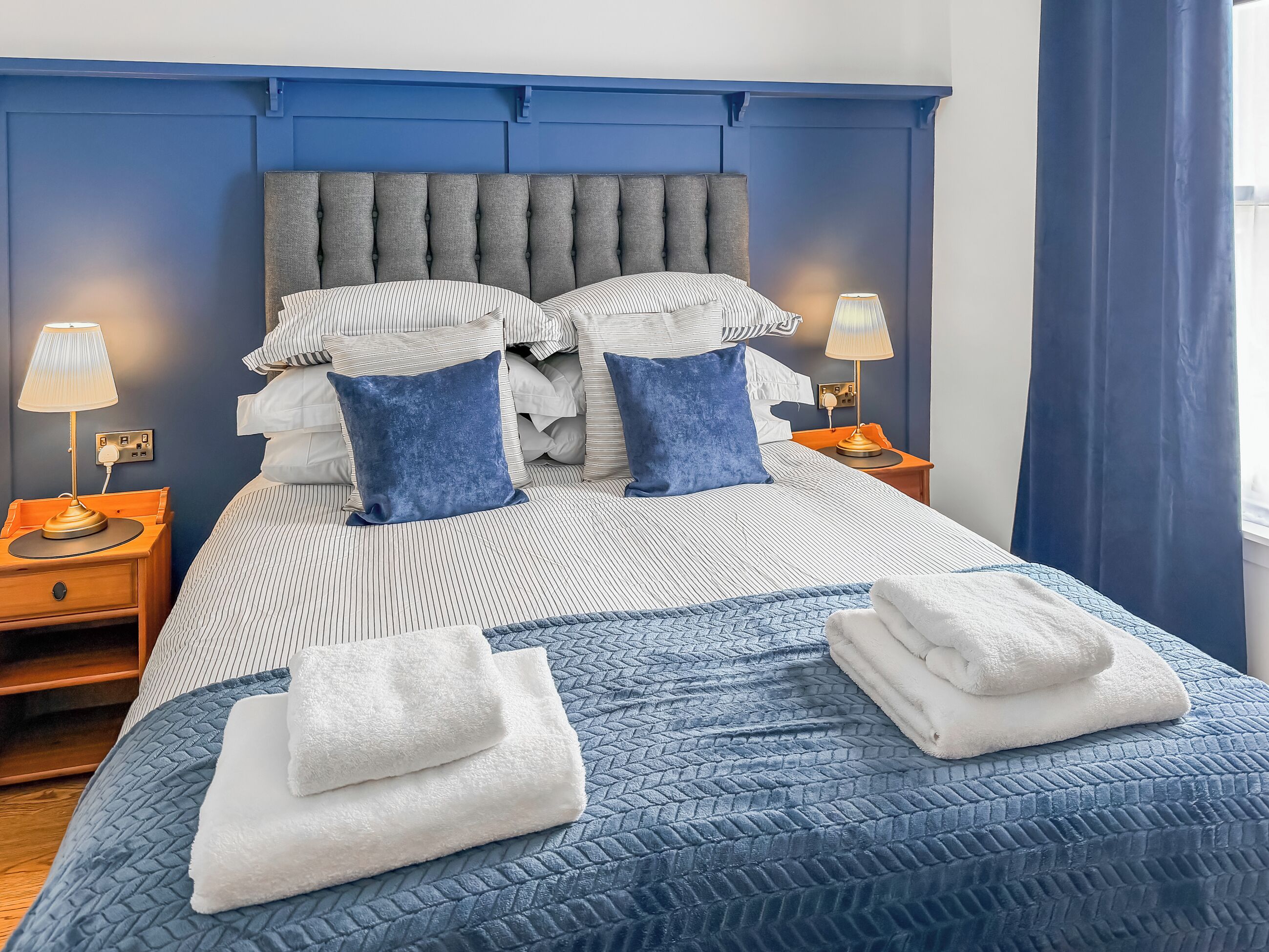 A hotel room with a large bed, two bedside tables, a blue headboard, and a window with curtains.