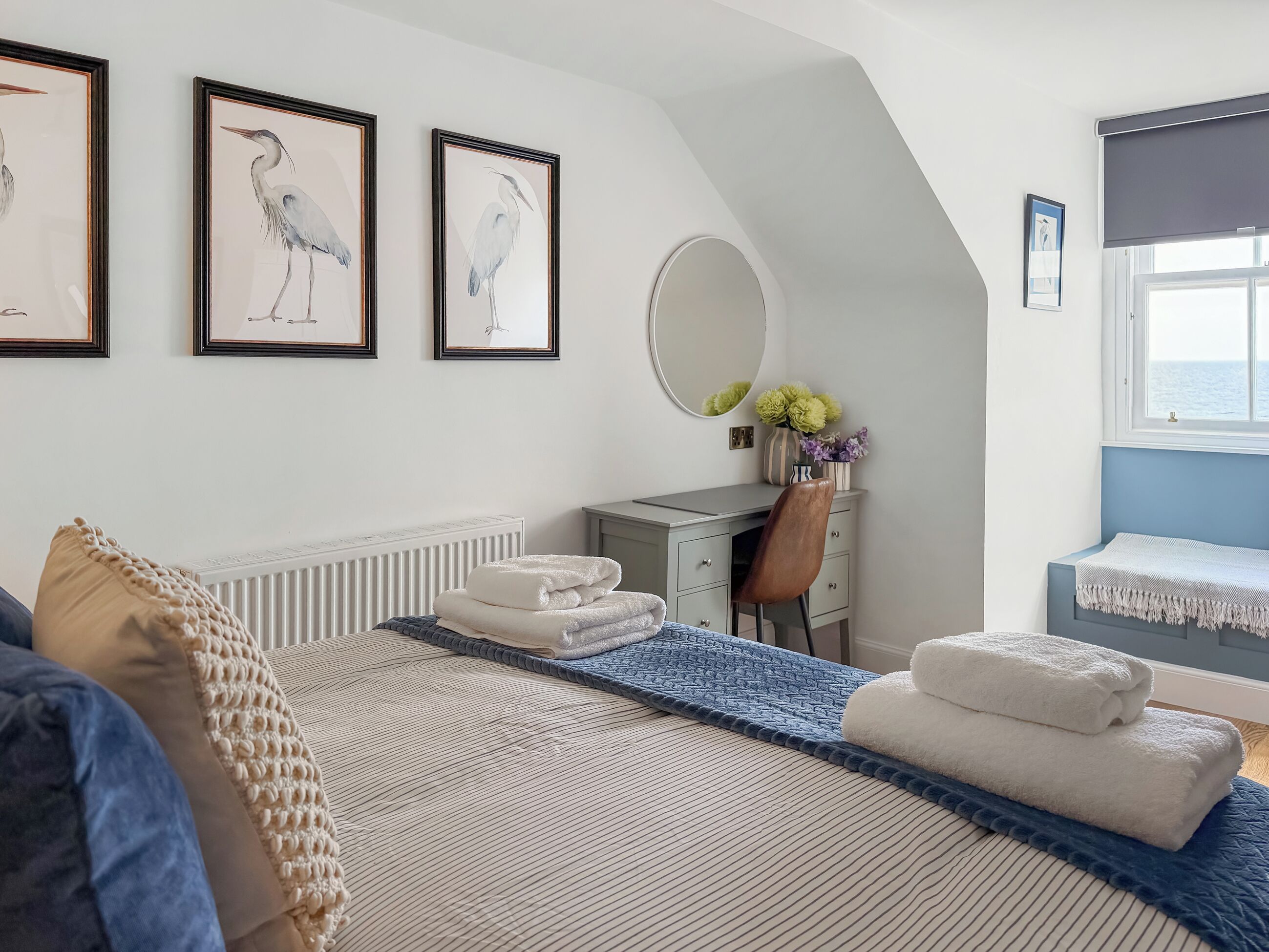 Un dormitorio con cama, escritorio, silla, radiador, ventana y cuadros en la pared.