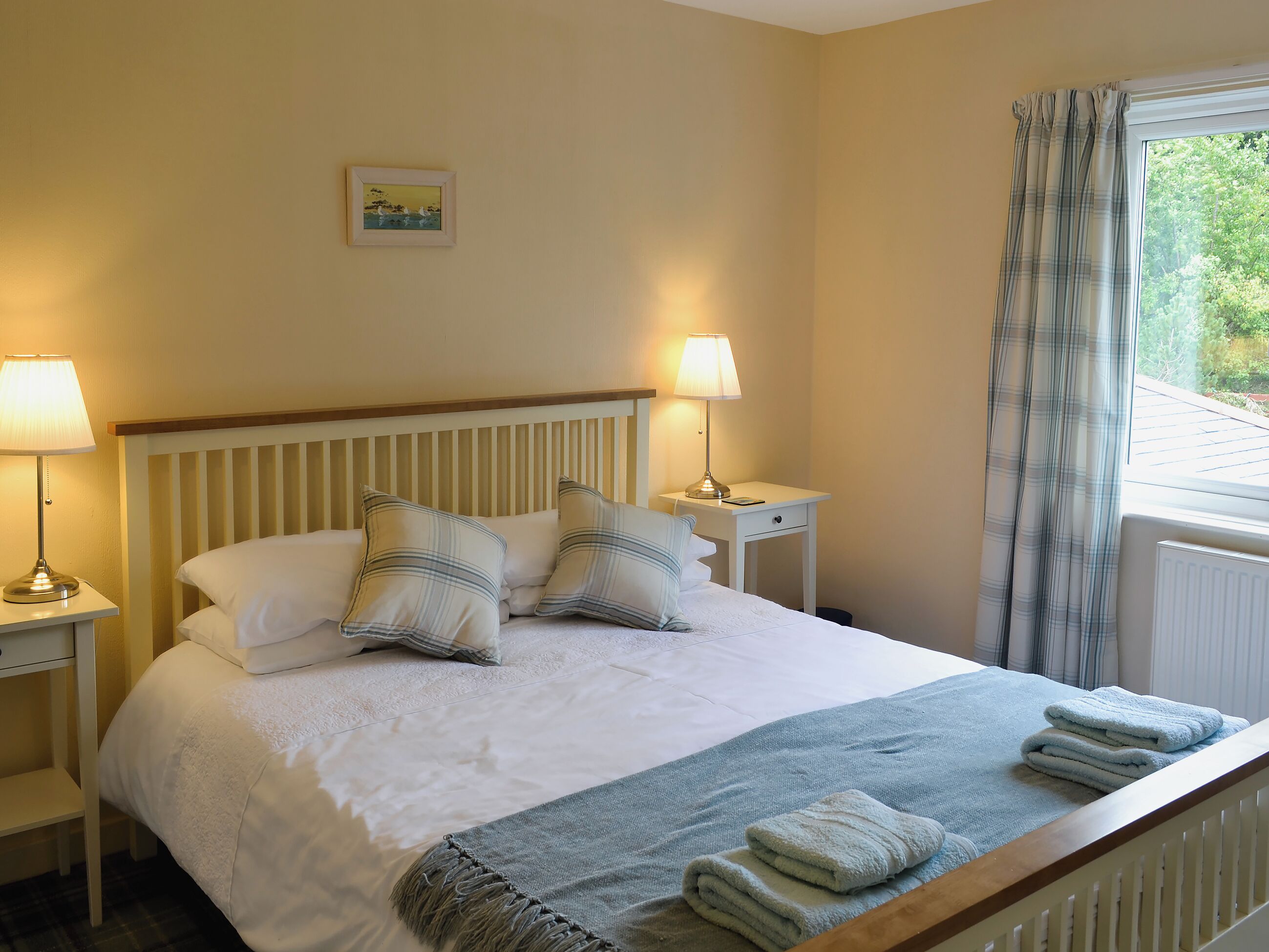 Een netjes opgemaakt bed met kussens, een nachtkastje met een lamp en een raam met gordijnen.