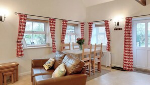 A living room with a brown leather sofa, wooden dining table, and chairs. Red curtains with white patterns hang on the windows. - 1 bedroom accommodation in Wansford, near Driffield (Wansford, near Driffield)