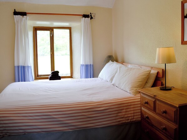 A bedroom with a bed, a nightstand, a lamp, a window with curtains, and a view of greenery outside. - Brothersfield Cottage (Hartsop, near Patterdale)