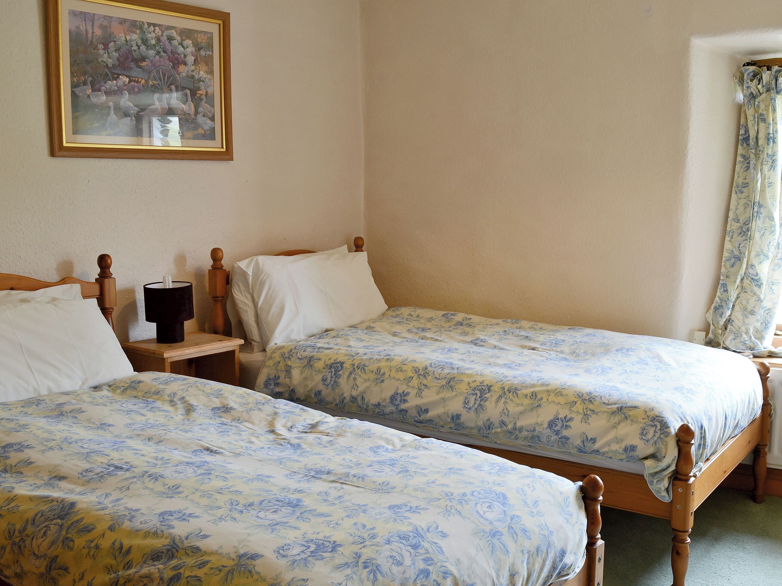 Ein Zimmer mit zwei Holzbetten, einem Nachttisch mit Lampe, einem gerahmten Bild an der Wand und einem Fenster mit Vorhängen.