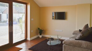 Un salon avec un téléviseur fixé au mur, une table basse en verre, un canapé, une plante en pot et une porte donnant sur un balcon.
