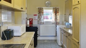 Une cuisine de taille réduite, équipée d’appareils électroménagers de base, avec une horloge au mur et une fenêtre agrémentée de rideaux à motifs floraux.