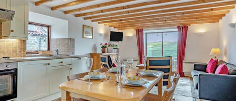 A cozy kitchen with wooden beams, a dining area with a table set for two, and a living area with a sofa and a TV.