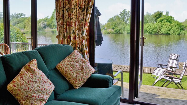 Een kamer met een groene bank, bloemkussens en uitzicht op een meer door grote ramen.