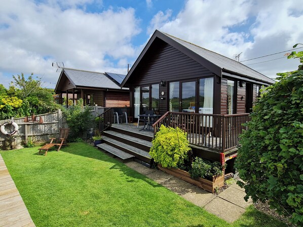 A wooden house with a deck, a small staircase leading to the deck, and a grassy yard.
