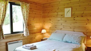 Un dormitorio de cabaña de madera con cama, almohadas, una mesita de noche, una lámpara y una ventana con cortinas.