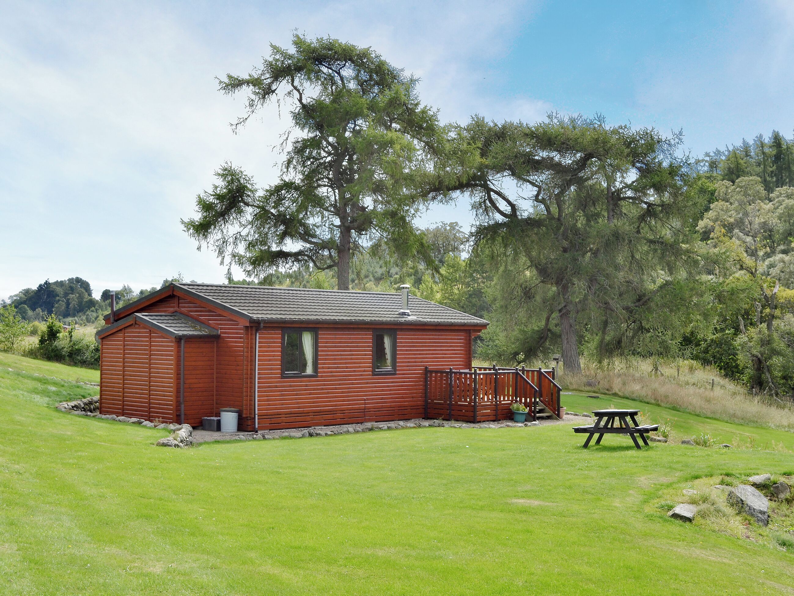 Une petite cabane en bois d’un seul étage, dotée d’une terrasse entourée d’une clôture et d’une table de pique-nique, située dans un espace verdoyant.