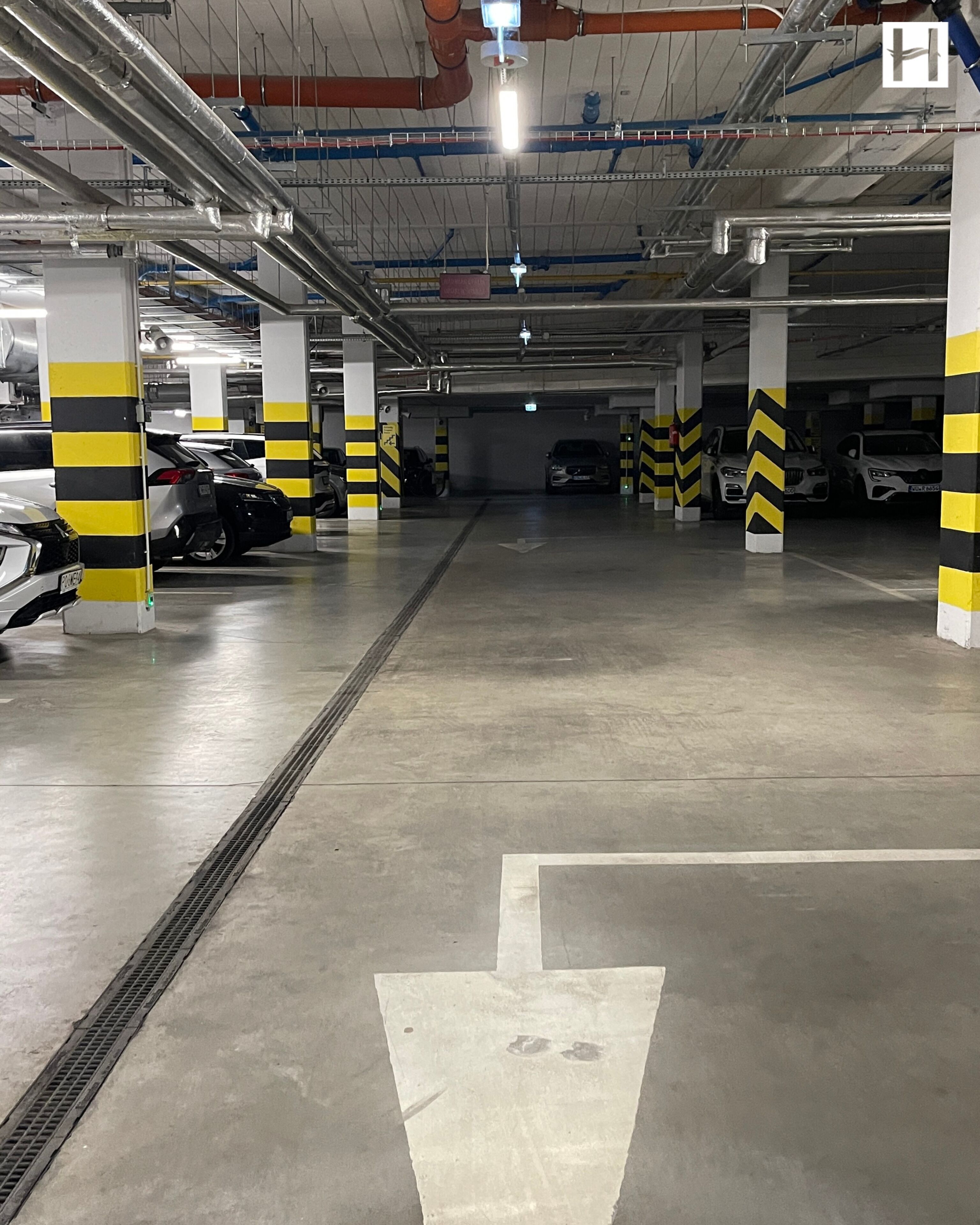 A multi-level parking garage with clear directional arrows on the floor, yellow and black safety barriers, and a ventilation system on the ceiling.