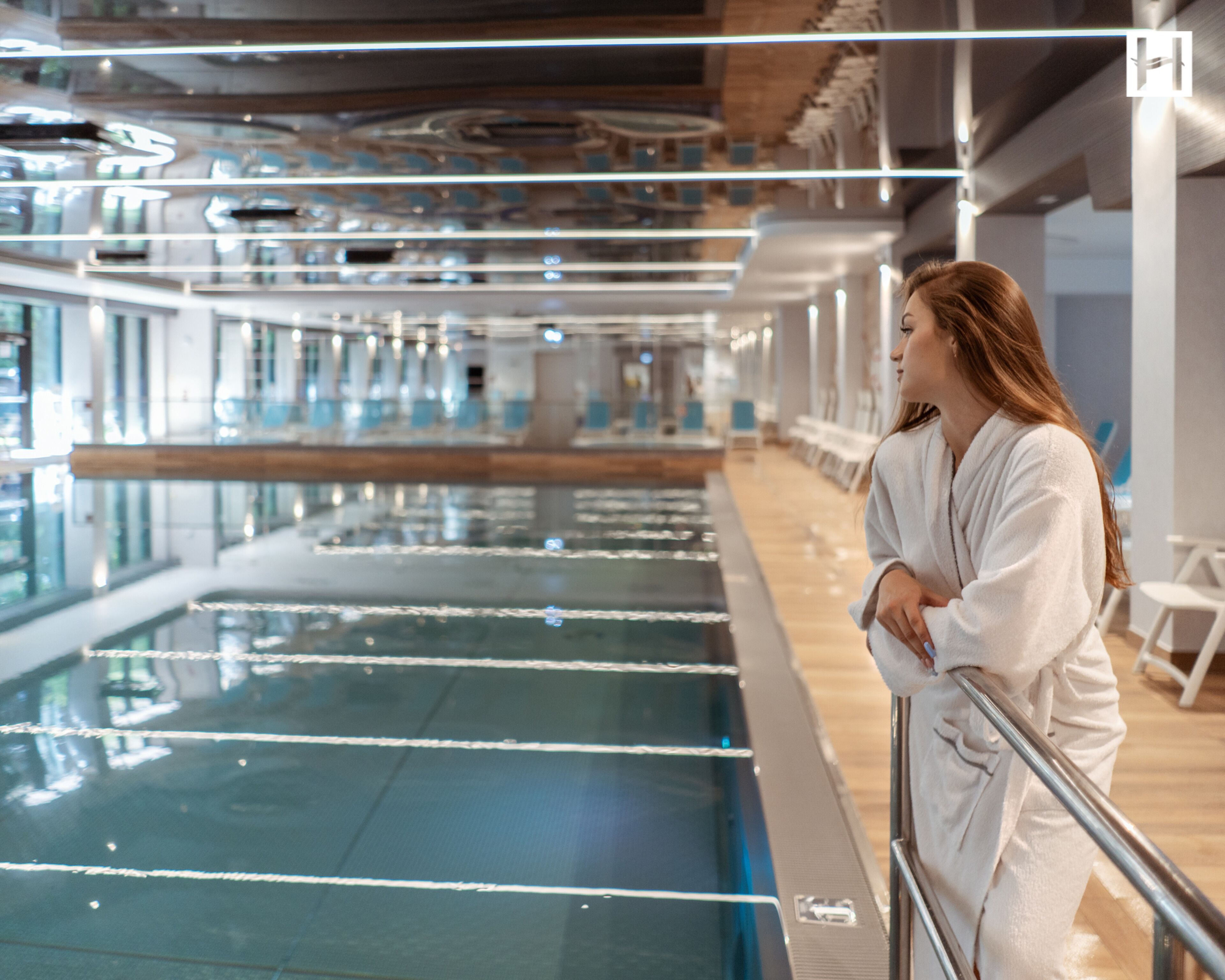 Una persona vestida con una bata blanca de pie junto a una piscina con barandilla, en una habitación con diseño moderno y ventanas amplias.