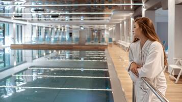 Una persona vestida con una bata blanca de pie junto a una piscina con barandilla, en una habitación con diseño moderno y ventanas amplias.