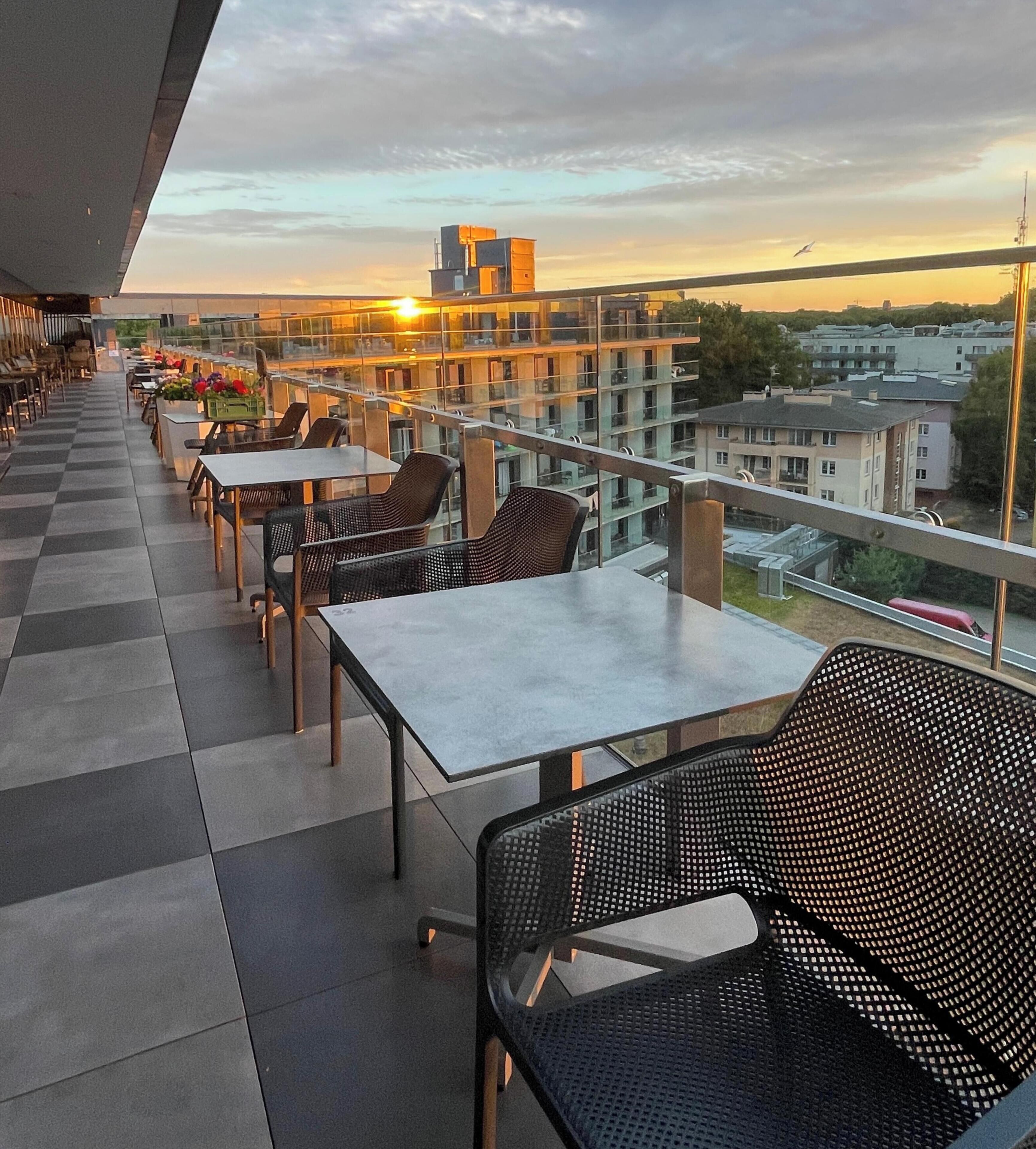 An outdoor terrace with tables and chairs, overlooking a cityscape during sunset.