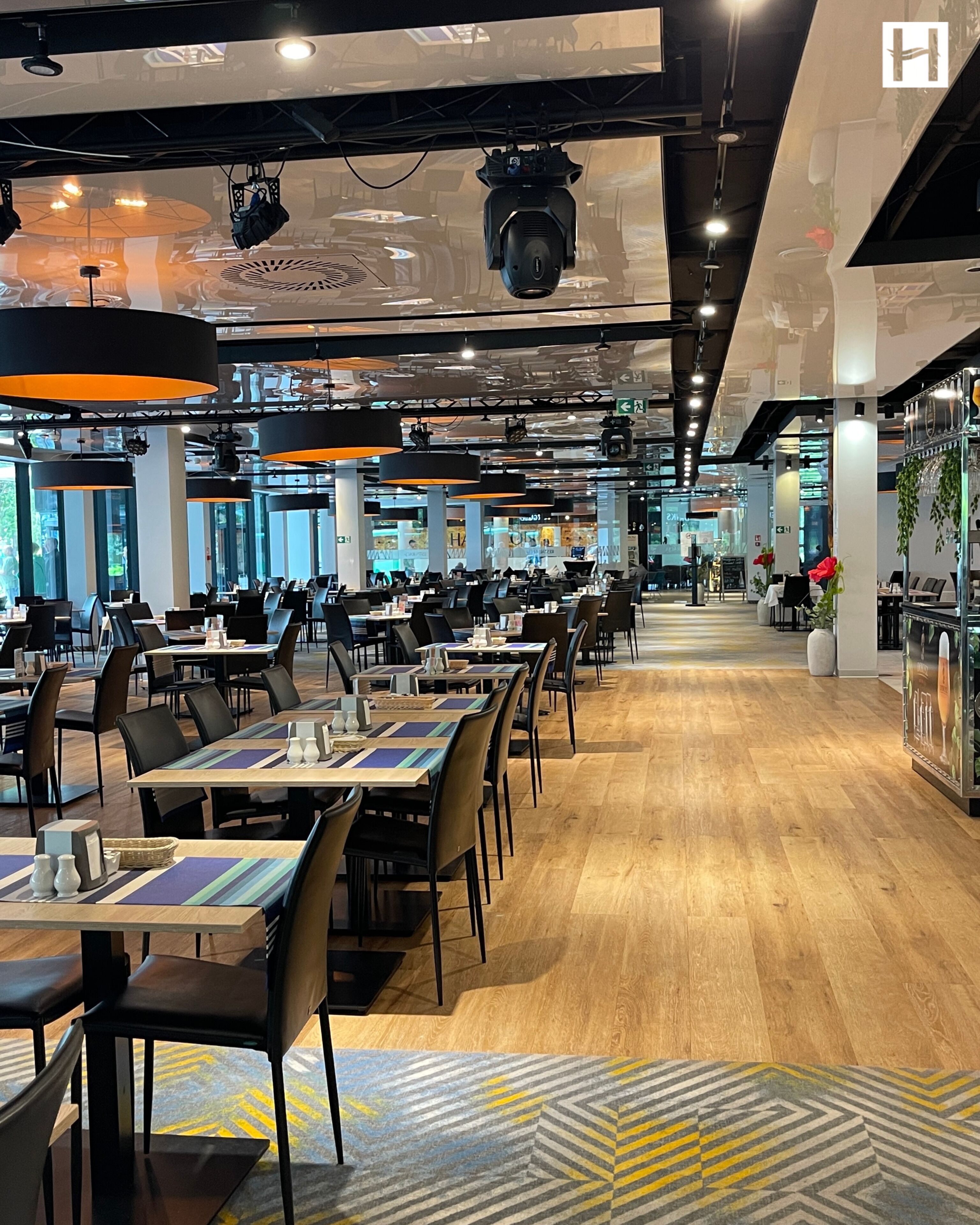 A spacious restaurant with multiple dining tables and chairs, overhead lighting, and a modern interior design.