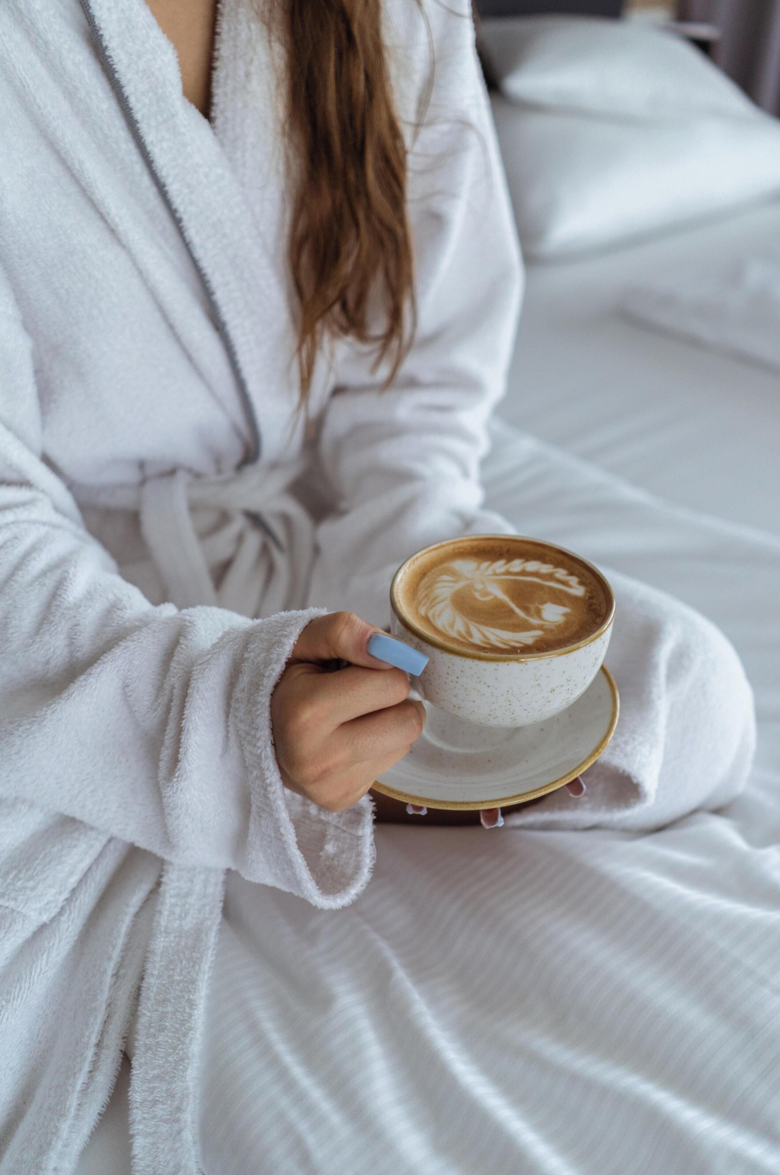 Een persoon in een witte badjas, koffie met latte-art vasthoudend.