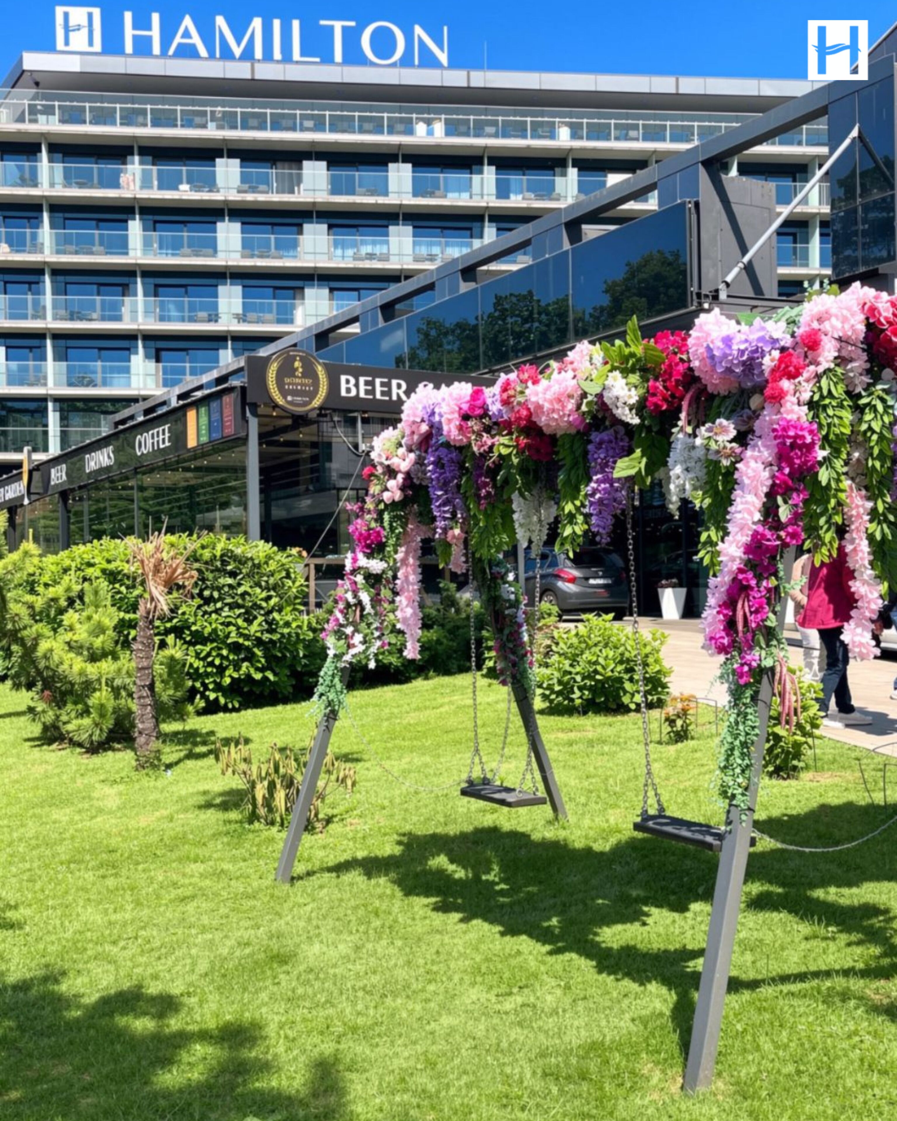 En hotellentrén med en gunga som är dekorerad med blommor, en skylt för ett café som serverar öl och kaffe, och en modern byggnad med en skylt som visar texten ”HAMILTON”.