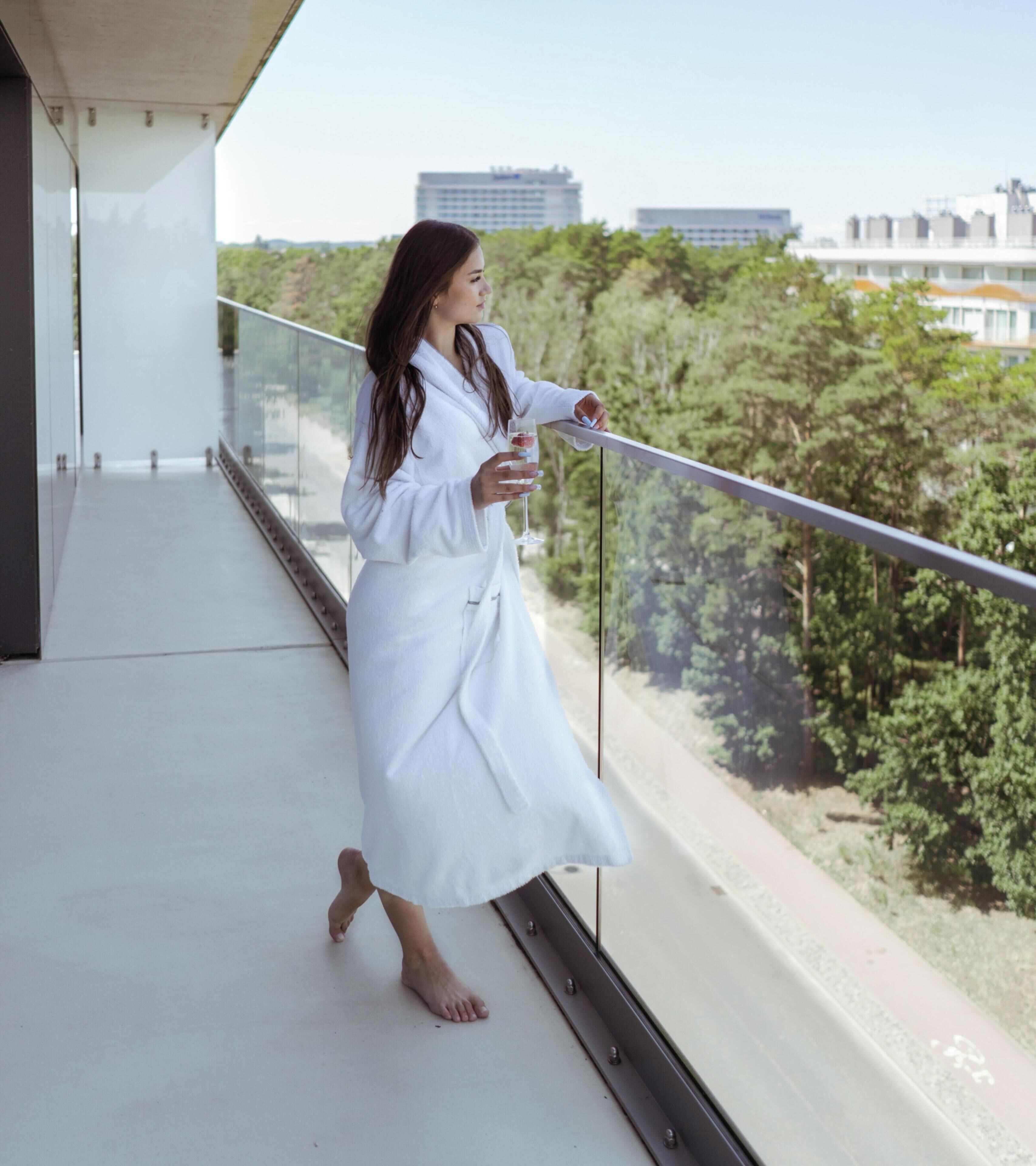 Une personne vêtue d’une longue robe blanche se tient sur un balcon doté d’une balustrade en verre, d’où elle domine un paysage urbain composé d’immeubles et d’arbres.