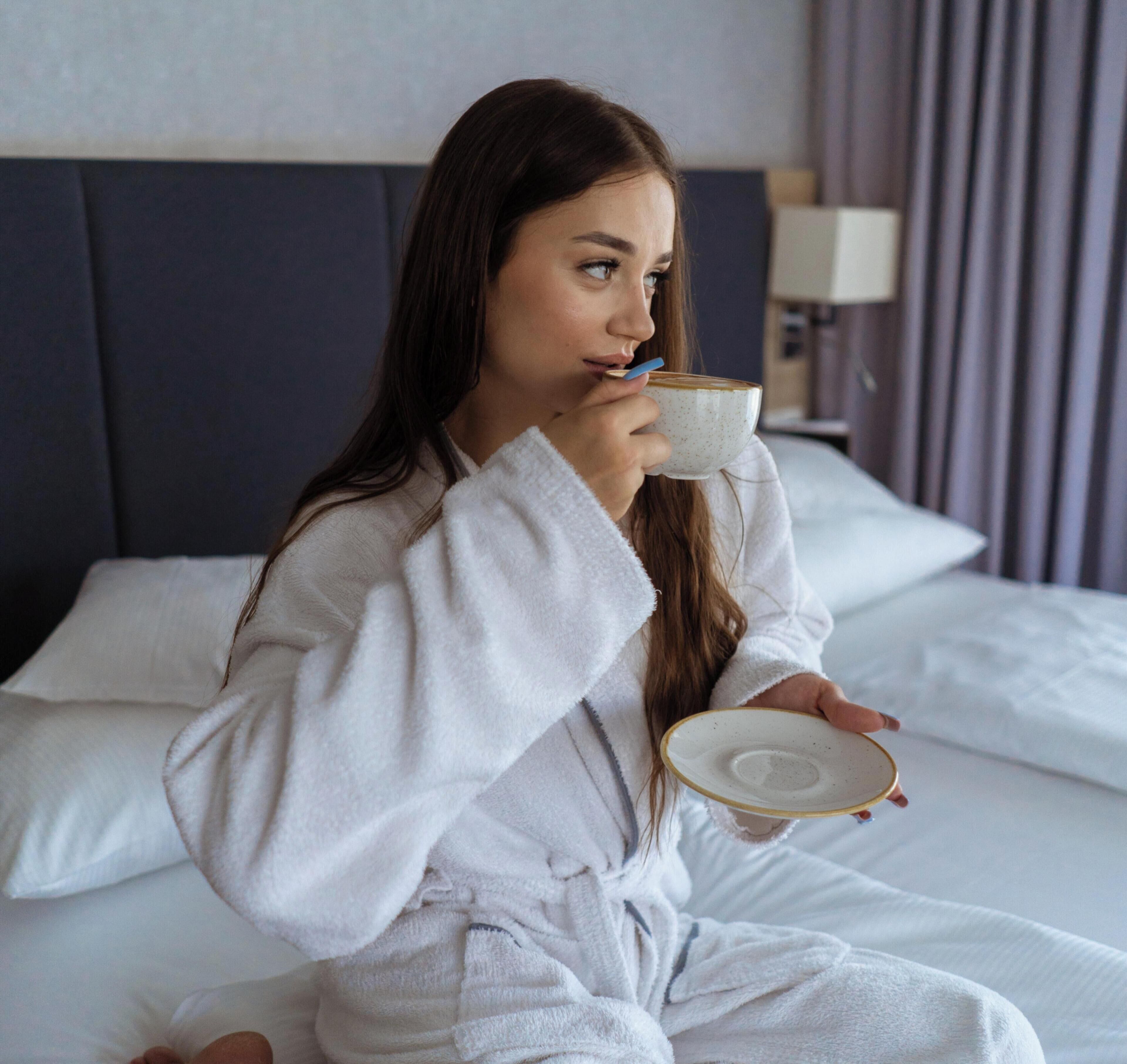 A person sitting on a bed in a hotel room, holding a cup and a plate.