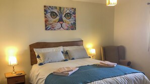 Een net opgemaakt bed met een blauwe deken, twee bedlampjes en een schilderij van een kattengezicht aan de muur.