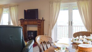 Een woonkamer met een televisie op een houten standaard, een bank, een eettafel met theeservies en uitzicht op een balkon door grote ramen.