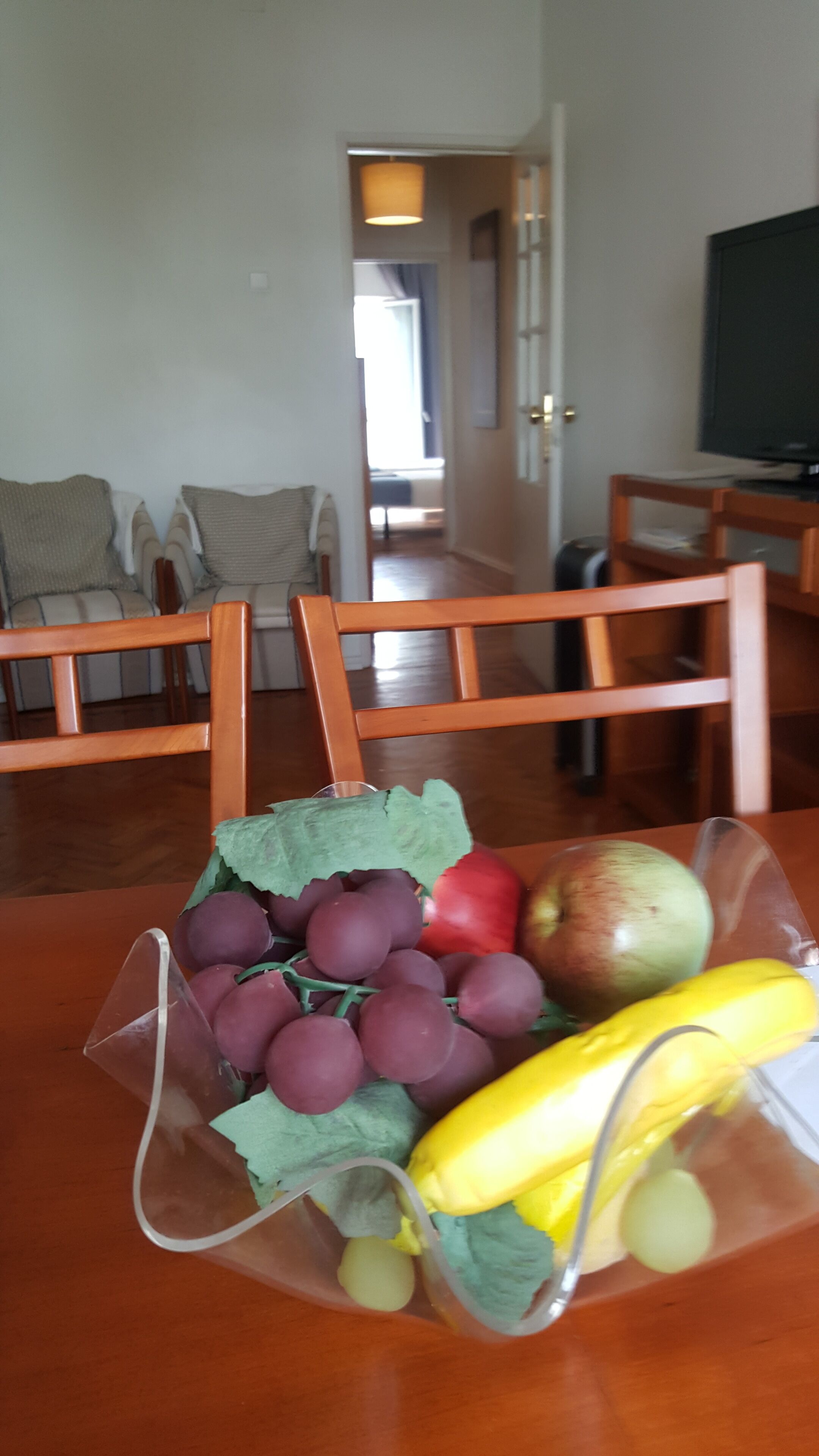 Ett matbord med en fruktskål som innehåller druvor, ett äpple, en banan och en melon. I förgrunden står en trästol och i bakgrunden syns en tv.