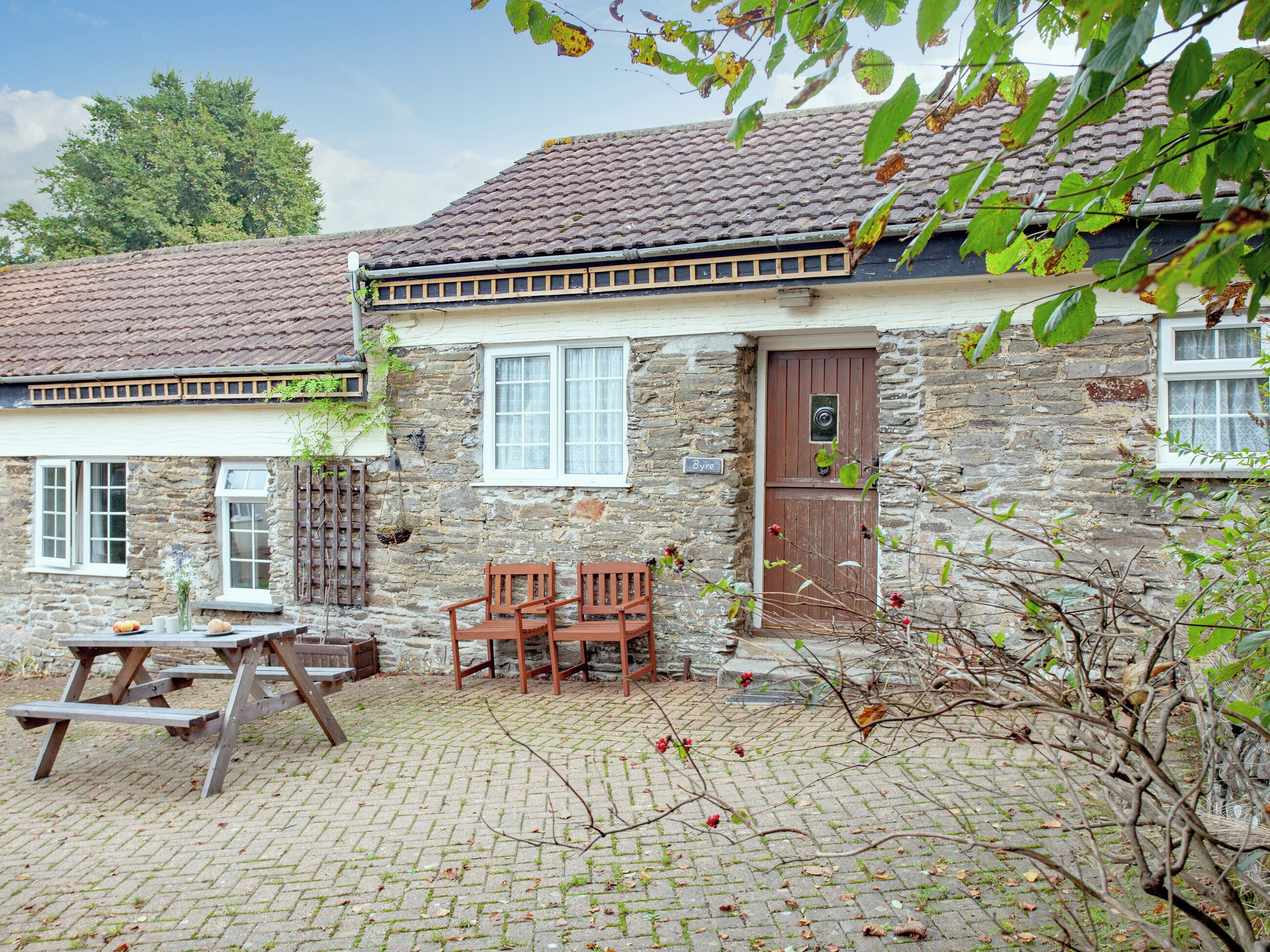Een stenen huis met pannendak, een houten deur en een kleine tuin met een picknicktafel en stoelen.
