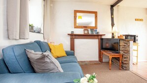 A living room with a blue sofa, a television on a stone fireplace, and a wooden table with a lamp. - 1 bedroom accommodation in Looe (Looe)