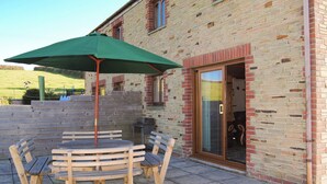 Une terrasse extérieure avec une table et des chaises en bois, un parasol vert et un bâtiment en briques doté d’une porte en verre.