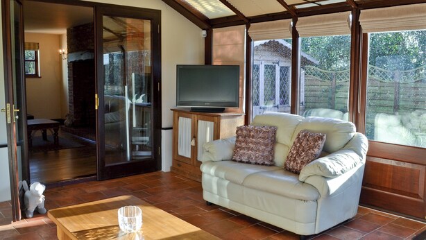 A living room with a sofa, a television on a stand, a glass door leading to a patio, and a tiled floor. - 4 bedroom accommodation in Dumpling Green, near Dereham (Dumpling Green, near Dereham)