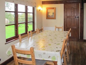 A dining room with a table covered in a leaf-patterned tablecloth, wooden chairs, a window with curtains, a wall-mounted light, and a framed picture. - 4 bedroom accommodation in Dumpling Green, near Dereham (Dumpling Green, near Dereham)