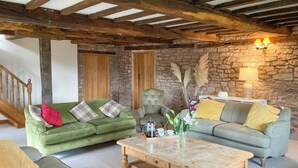 Un salon chaleureux avec un mur en pierre, des poutres en bois, deux canapés verts ornés de coussins à motifs, une table basse en bois et un escalier en arrière-plan.