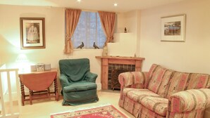A living room with a green armchair, a patterned sofa, a red floral rug, a fireplace, and two framed pictures on the walls.
