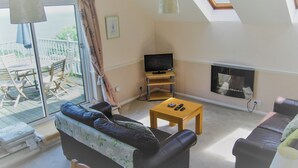 A living room with a sofa, a television, a fireplace, and a balcony with outdoor furniture. - 2 bedroom accommodation in Maidencombe, near Torquay (Maidencombe, near Torquay)