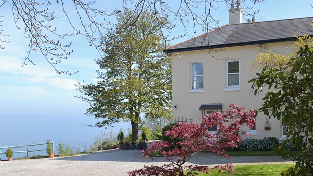 A house with a gravel driveway, a large tree, and a view of the sea. - 2 bedroom accommodation in Maidencombe, near Torquay (Maidencombe, near Torquay)