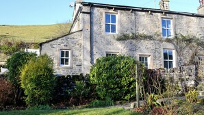 A stone house with three windows, surrounded by greenery and a grassy hill in the background.
