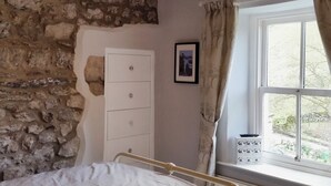 A bedroom with a stone wall, a bed, a window with curtains, and a bedside table with a lamp.