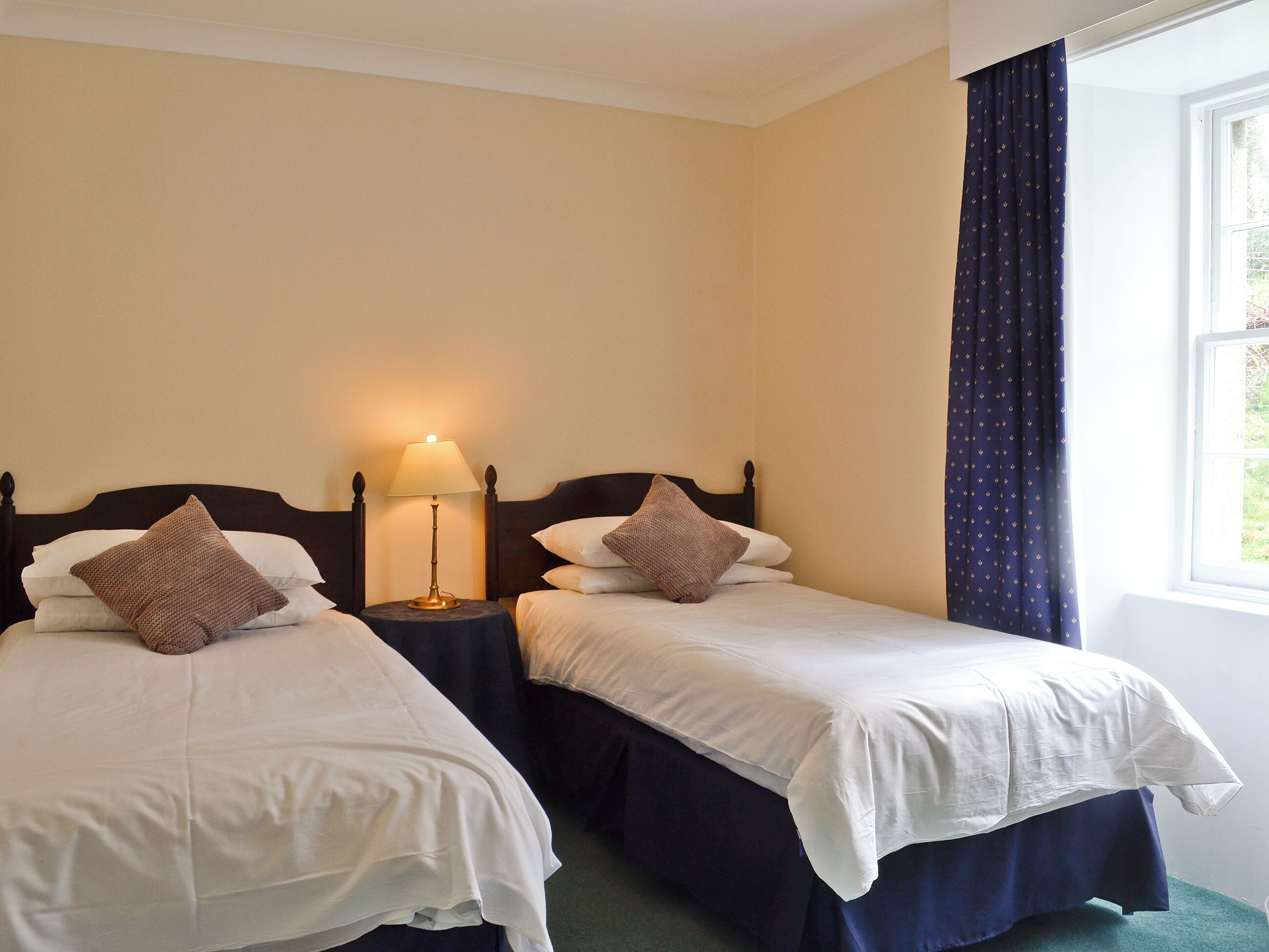 A hotel room with two single beds, a bedside table with a lamp, and a window with curtains.