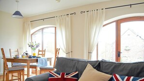 A living room with a grey sofa, two British flag cushions, a wooden dining table with chairs, and a window with curtains.