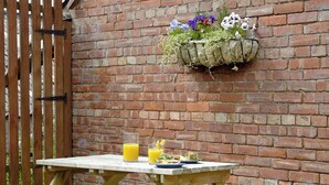 A wooden picnic table with benches, a hanging flower planter, and a brick wall in the background.
