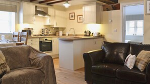 Un salon chaleureux avec un canapé et un divan en cuir, une cuisine dotée de meubles blancs et d’un îlot central intégré, et des poutres en bois au plafond.